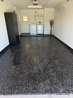 A room with a speckled black floor, a black refrigerator, two white appliances, and a broom in the corner.