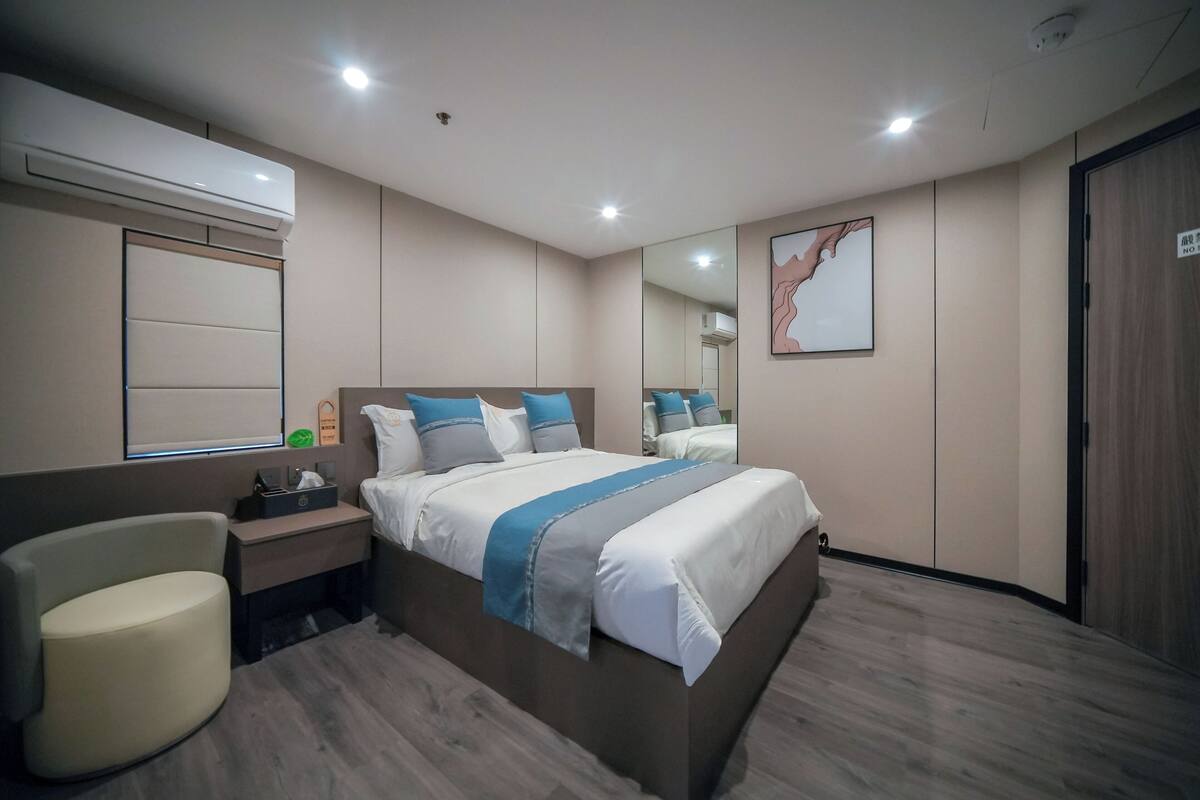 A modern hotel room with a large bed, bedside tables, a chair, a mirror, and an air conditioning unit.