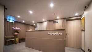 Zona de recepción con un mostrador de madera identificado con la etiqueta "Reception", un pequeño rincón de estar con una silla y una mesa, y una orquídea en maceta.
