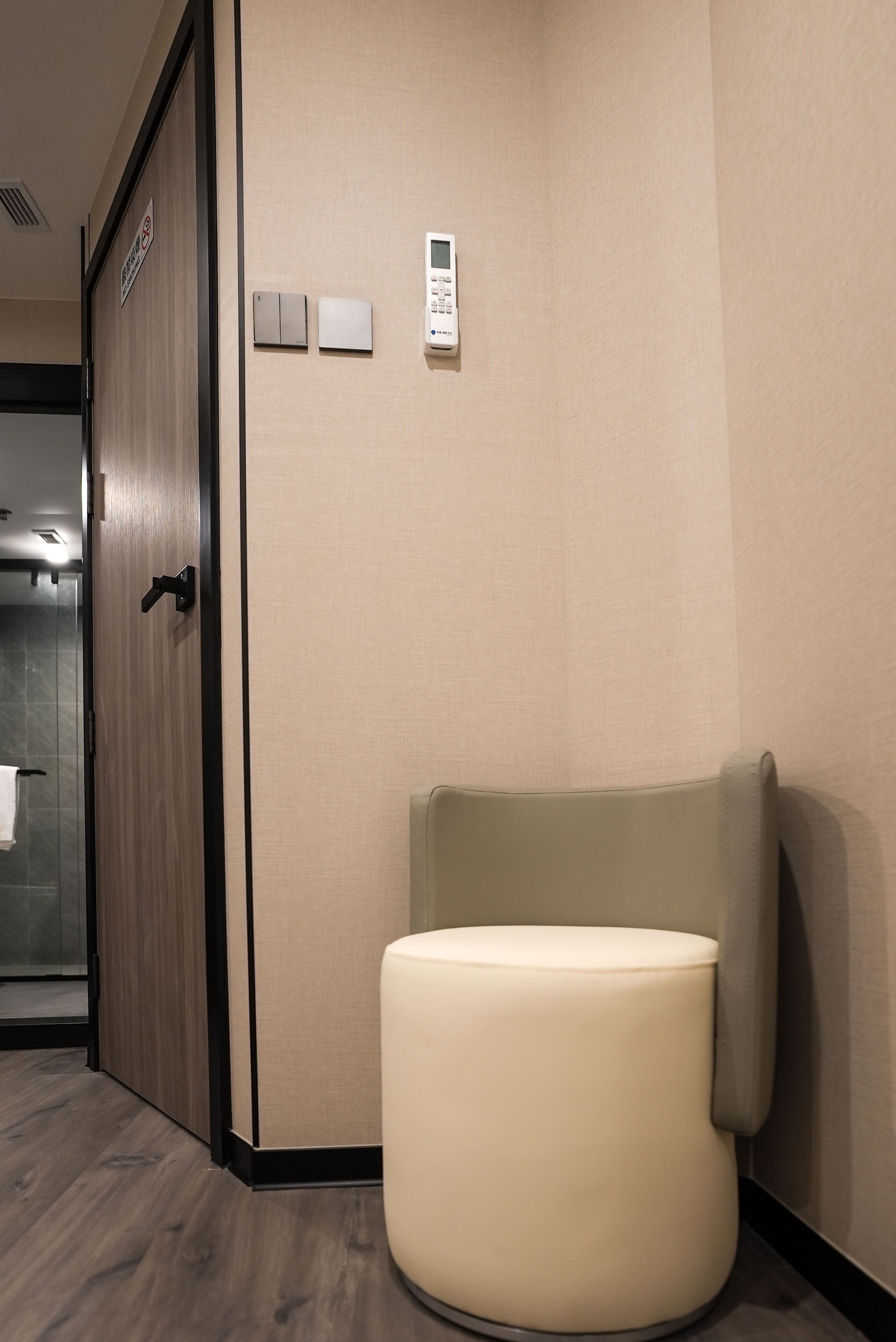 A hotel room with a wooden door, a small white control panel on the wall, and a round white ottoman.