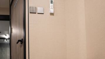 A hotel room with a wooden door, a small white control panel on the wall, and a round white ottoman.