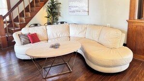 A living room with a L-shaped sectional sofa, a round coffee table, a wooden staircase, and a potted plant. - Charming Historic 5BR Home • Close to Universities, Hospital & Downtown (Flint)
