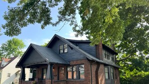 A two-story house with a brick exterior, a dark roof, and a front porch. - Charming Historic 5BR Home • Close to Universities, Hospital & Downtown (Flint)