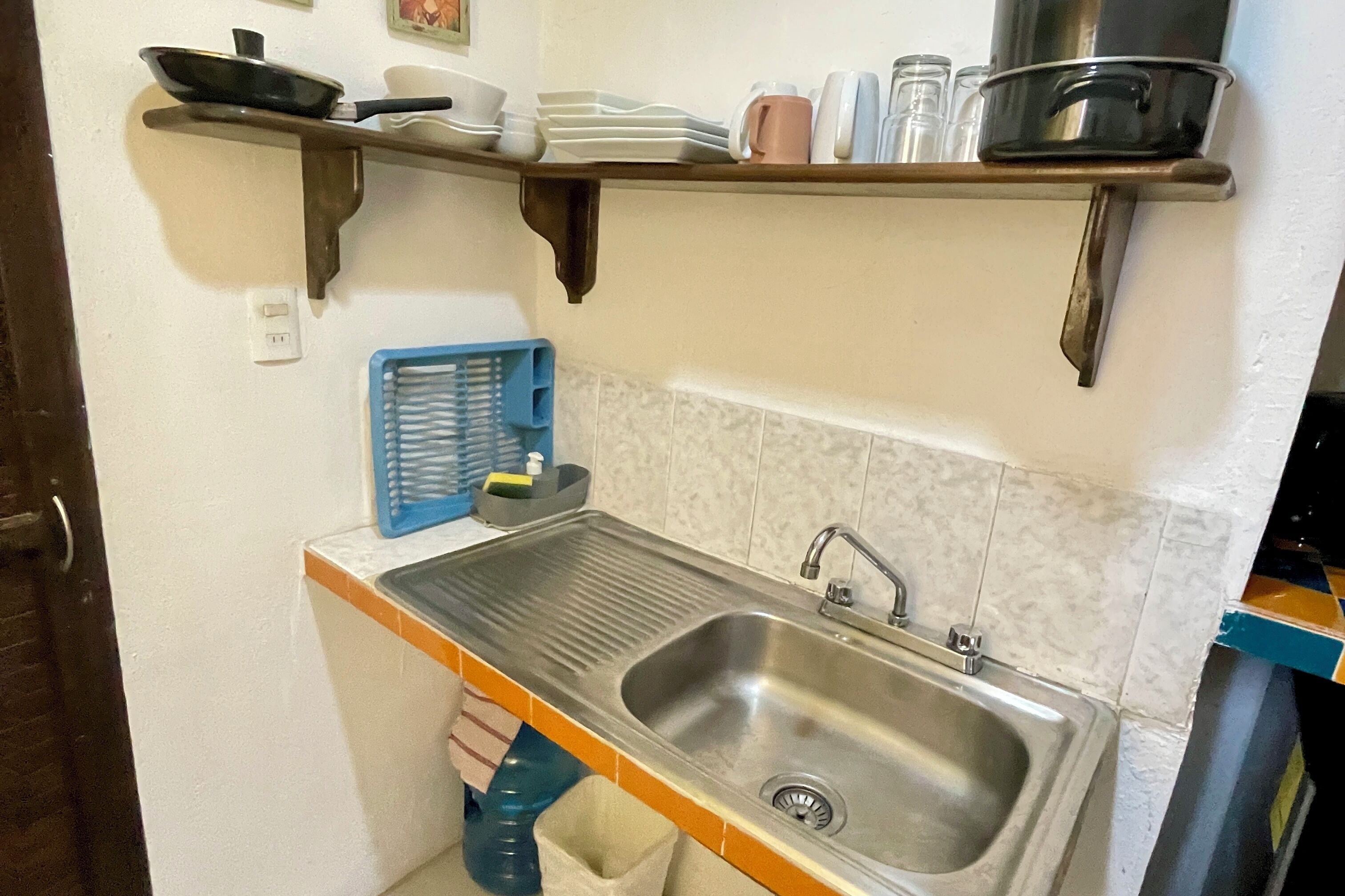 A kitchen with a stainless steel sink, a blue dish rack, and a shelf above holding dishes and a pot.
