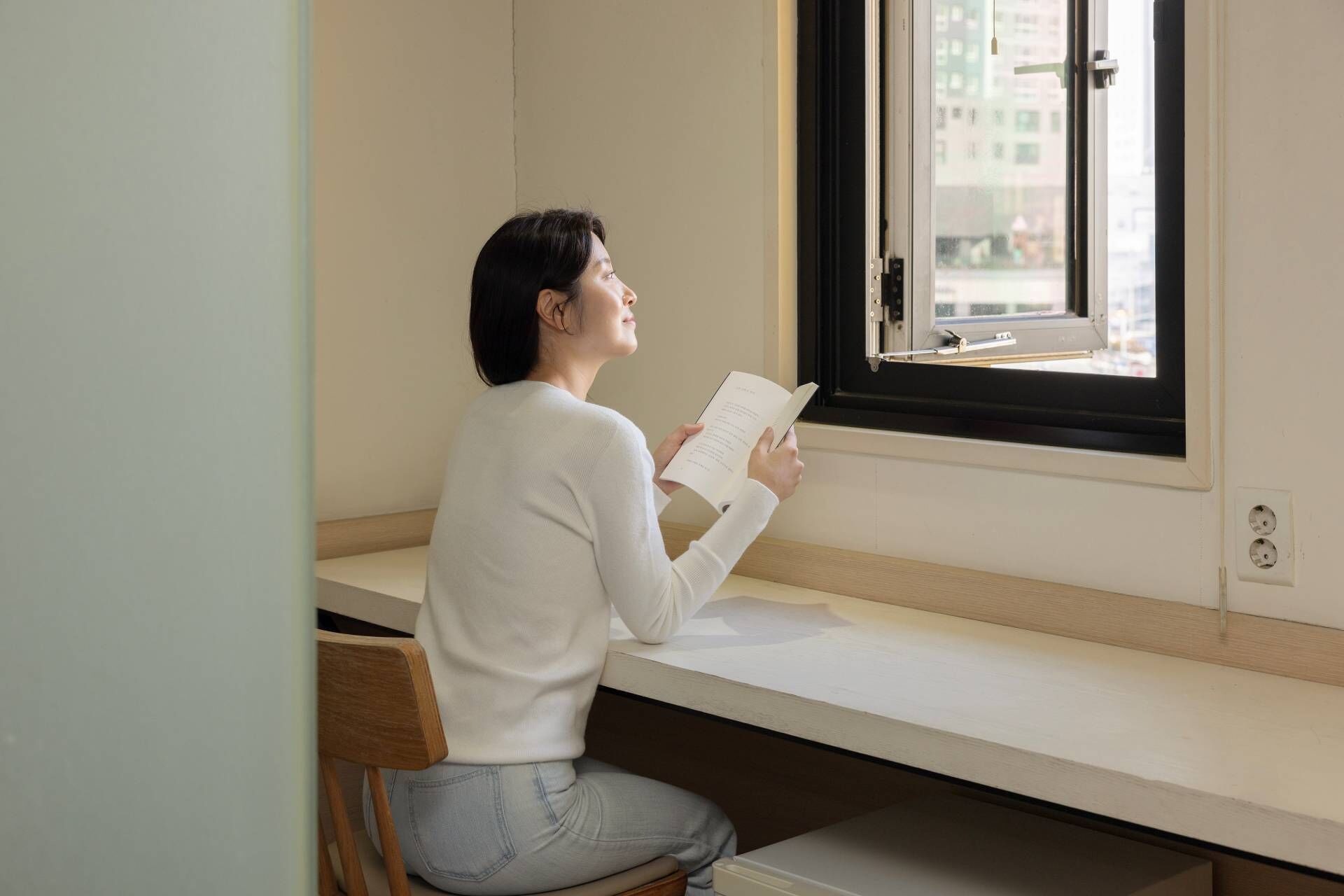 A person sitting on a bench by a window, reading a book.