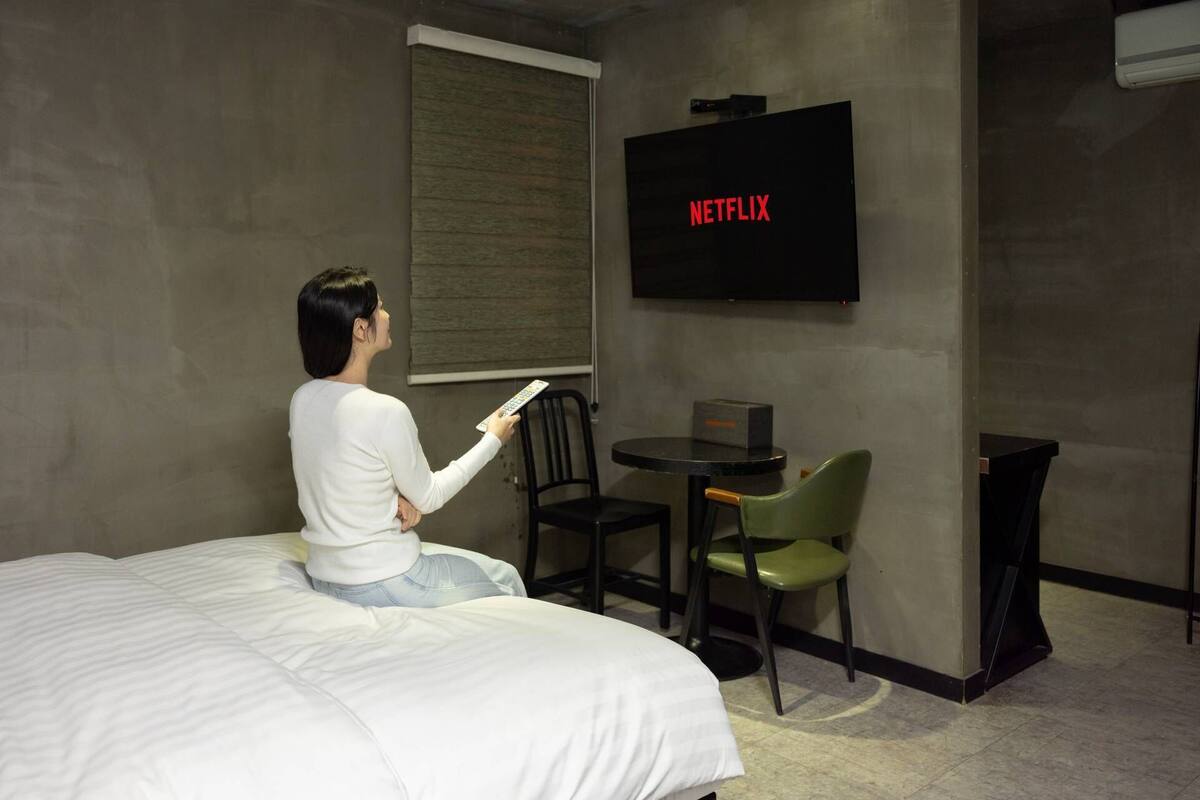 A hotel room with a bed, a TV mounted on the wall displaying 'NETFLIX', a small round table with a chair, and a window with blinds.