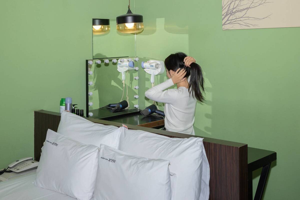 A hotel room with a green wall, a vanity with a mirror, and a hairdryer on the counter. There are white pillows with text on them and a telephone on the side table.