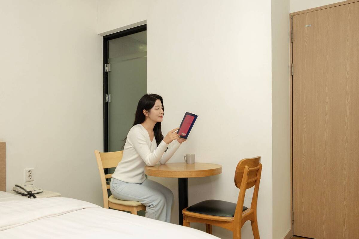 A hotel room with a bed, a desk, a chair, a wooden door, and a wall-mounted phone.
