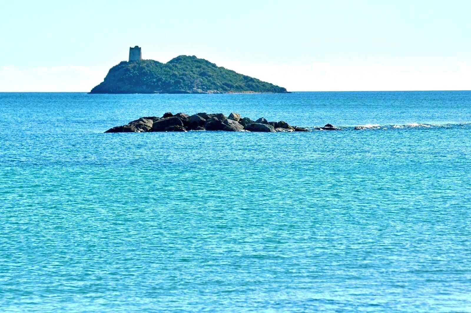 A clear view of a rocky island with a tower in the distance across a calm sea.