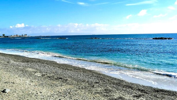 Una playa con aguas azul turquesa, orilla pedregosa y edificios a lo lejos.