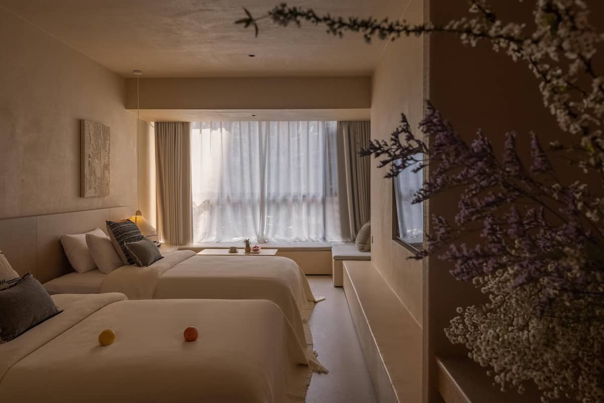 Et hotelværelse med to senge, et vindue med lette gardiner, et lille bord og en dekorativ plante.