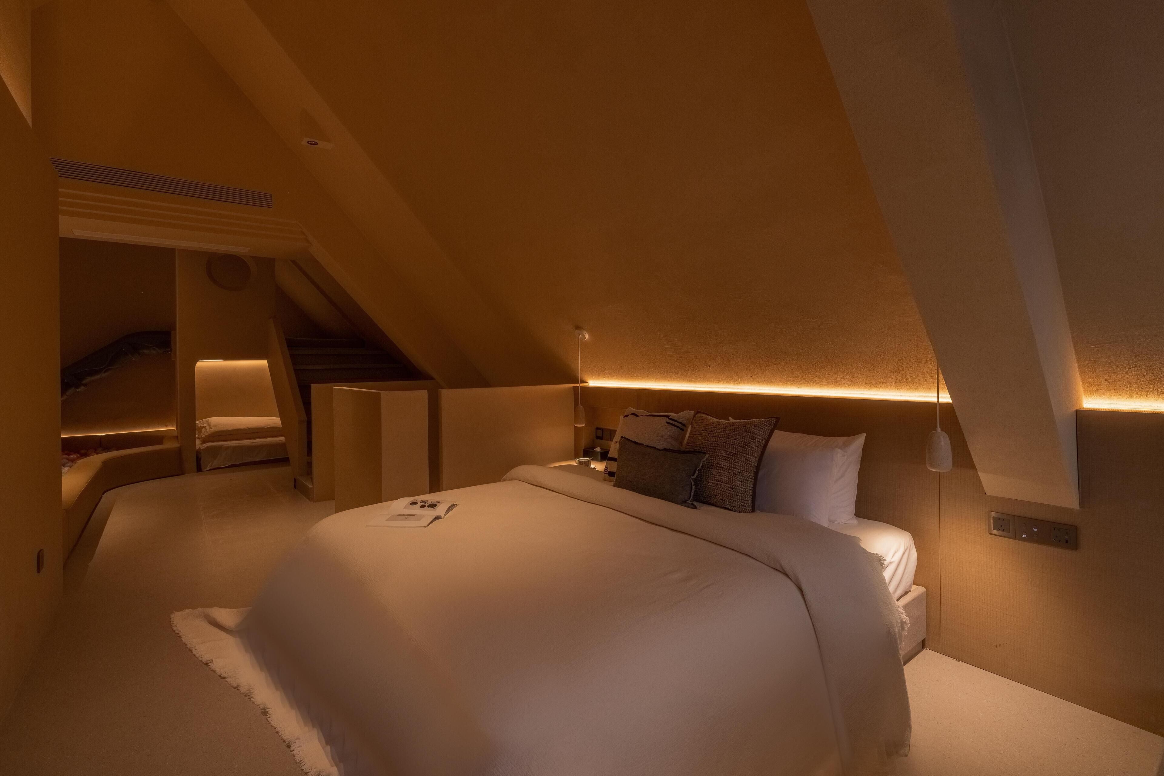 Un dormitorio moderno, con poca luz, que cuenta con una cama grande, una pequeña zona de estar y un techo inclinado.