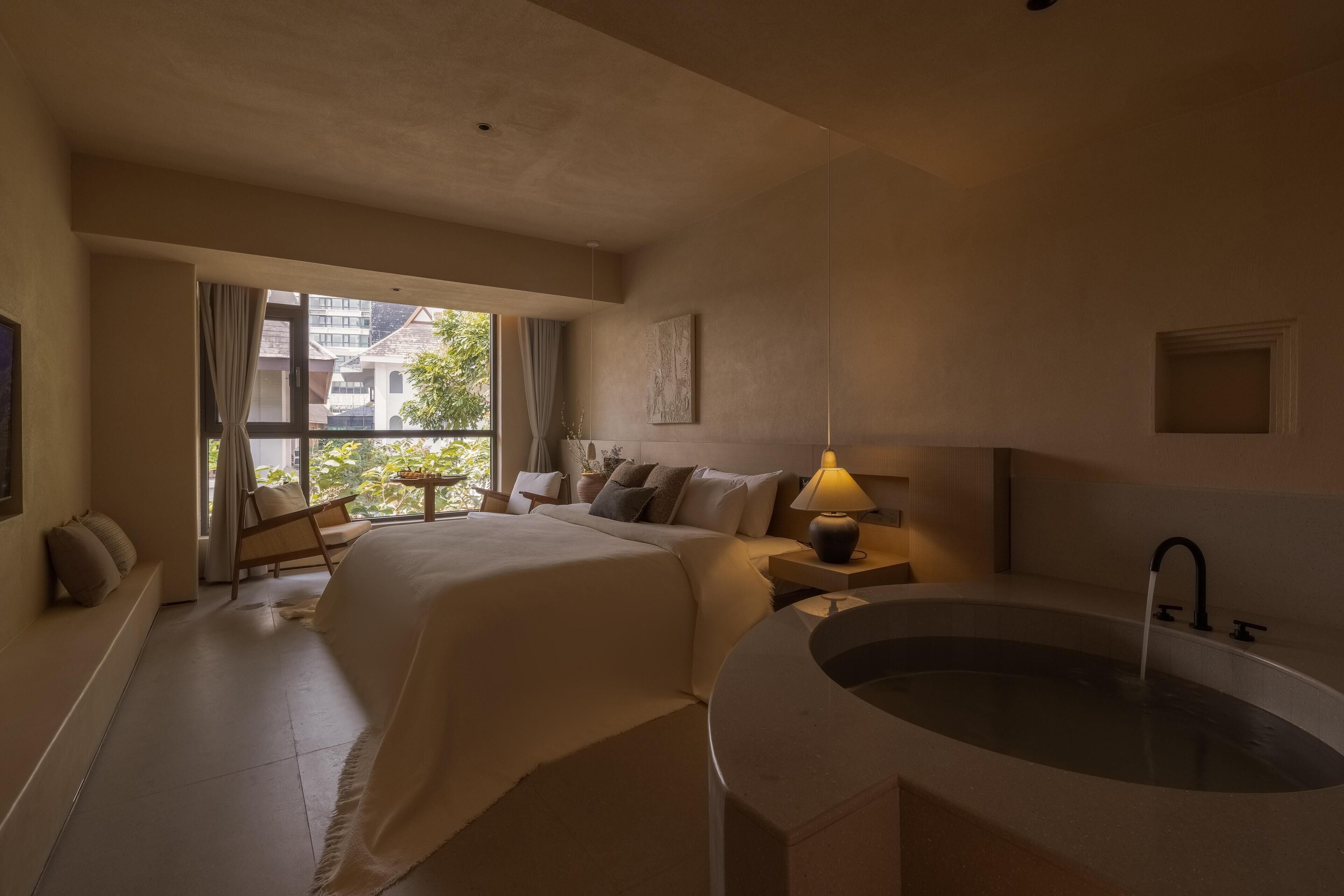 A spacious bedroom with a large bed, a sitting area, a bathtub, and a view of buildings through the window.
