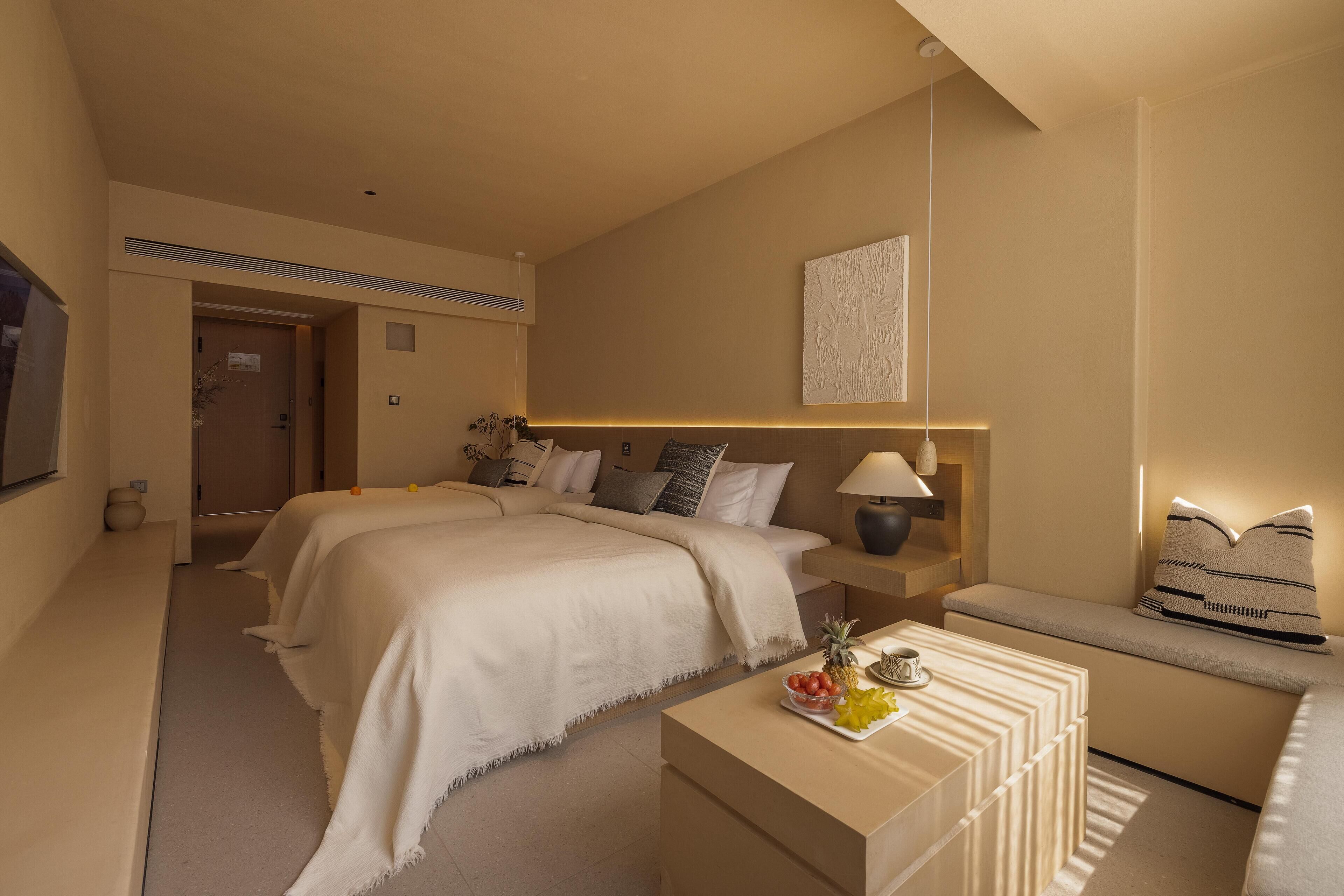 A hotel room with two beds, a bedside table with a lamp, a bench with cushions, and a small table with fruit.