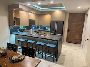A modern kitchen with a central island, bar stools, a dining table set for four, and a wooden door. - 1-bedroom beachfront condo in Playa Encanto Encantame Soleil (Puerto Penasco)