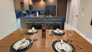 Zona pranzo con tavolo in legno apparecchiato per quattro persone, cucina sullo sfondo e moderno design del soffitto.