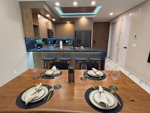 A dining area with a wooden table set for four, a kitchen in the background, and a modern ceiling design. - 1-bedroom beachfront condo in Playa Encanto Encantame Soleil (Puerto Penasco)