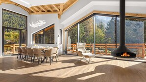 Une pièce spacieuse comprenant une grande table à manger et des chaises, une cheminée et de grandes fenêtres donnant sur une montagne boisée.