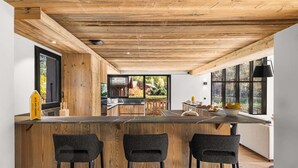 Eine moderne Küche mit Holzbalken, einer zentralen Kochinsel mit Barhocker und großen Fenstern mit Blick ins Freie.