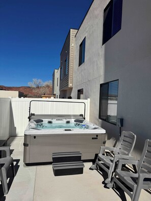 Ein Whirlpool mit Stufen, umgeben von Terrassenstühlen, in einer Außenanlage mit weißem Zaun und einem Gebäude im Hintergrund.