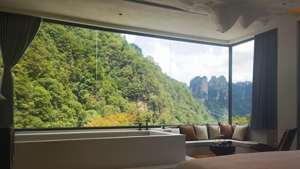 Ein modernes Schlafzimmer mit einem großen Fenster, einer Badewanne und Blick auf einen bewaldeten Berg.