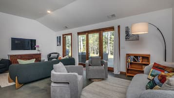 Un salon moderne avec un canapé gris, des fauteuils, une bibliothèque en bois et une grande fenêtre donnant sur la forêt.