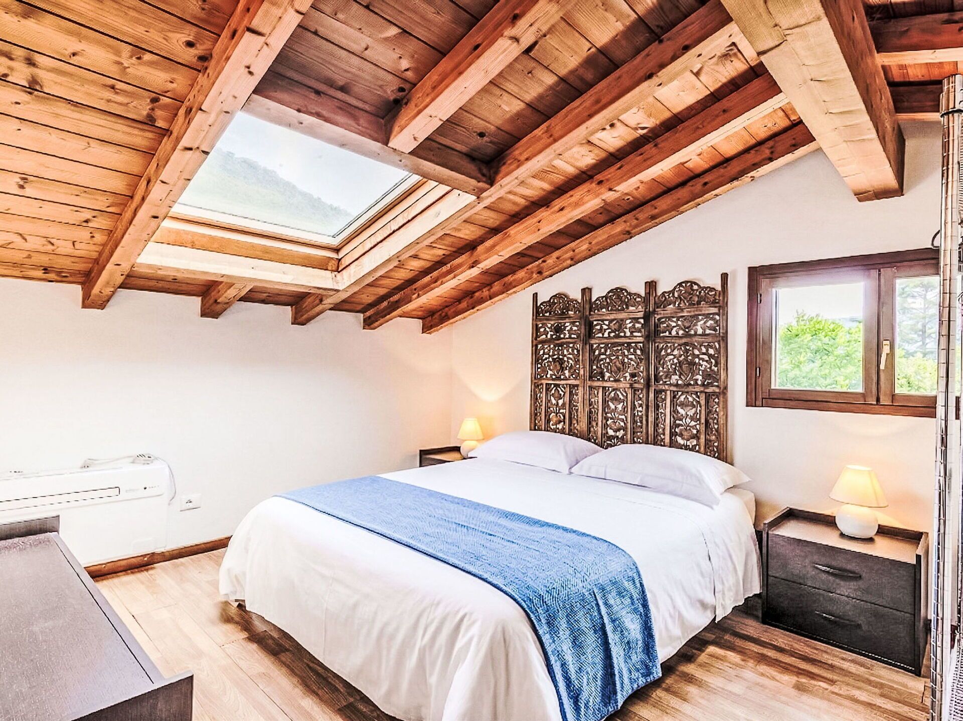 A bedroom with a skylight, wooden ceiling beams, a bed with white bedding and a blue throw, a nightstand with a lamp, and a window with a view of greenery.