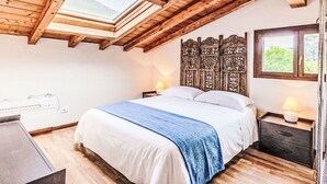 Un dormitorio con tragaluz, vigas de madera en el techo, una cama con ropa blanca y un plaid azul, una mesita de noche con lámpara, y una ventana con vistas a la vegetación.