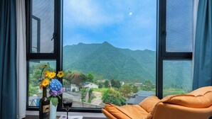 Een kamer met een groot raam met uitzicht op bergen, een houten tafel met een fles en bloemen, en een oranje stoel.