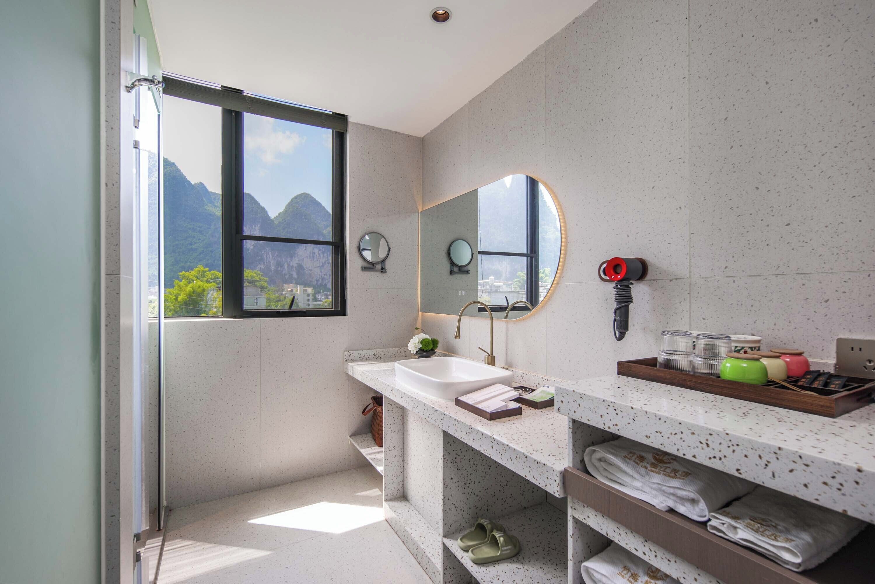 A modern bathroom with a large window, a sink, a mirror, and a shelf with towels and toiletries.
