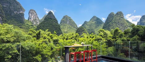 A glass-enclosed terrace with a table and red chairs, overlooking a lush forest and karst mountains.