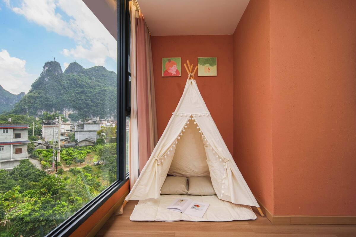 A room with a large window overlooking a mountainous landscape, a white tent-like bed with pillows, and two framed pictures on the wall.