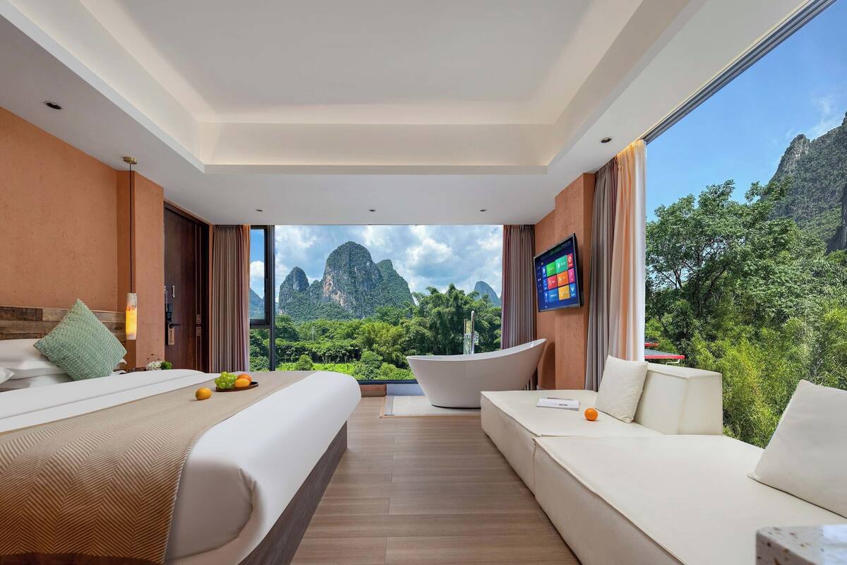 A modern hotel room with a large bed, a sofa, and a view of mountains and greenery.