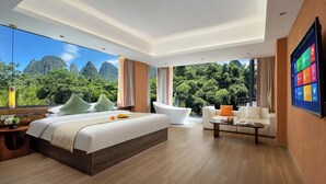 A modern hotel room with a large bed, a sitting area with a sofa and armchairs, a large window with a view of mountains and greenery, and a flat-screen TV mounted on the wall. - Riverside Bamboo Frest Hotel (Guilin)