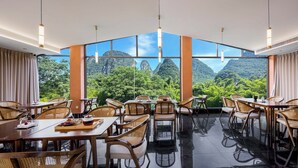 A dining area with tables and chairs, large windows, and a view of mountains. - Riverside Bamboo Frest Hotel (Guilin)