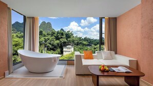 A modern hotel room with a large bathtub, a white sofa, and a coffee table with fruits and a book. - Riverside Bamboo Frest Hotel (Guilin)