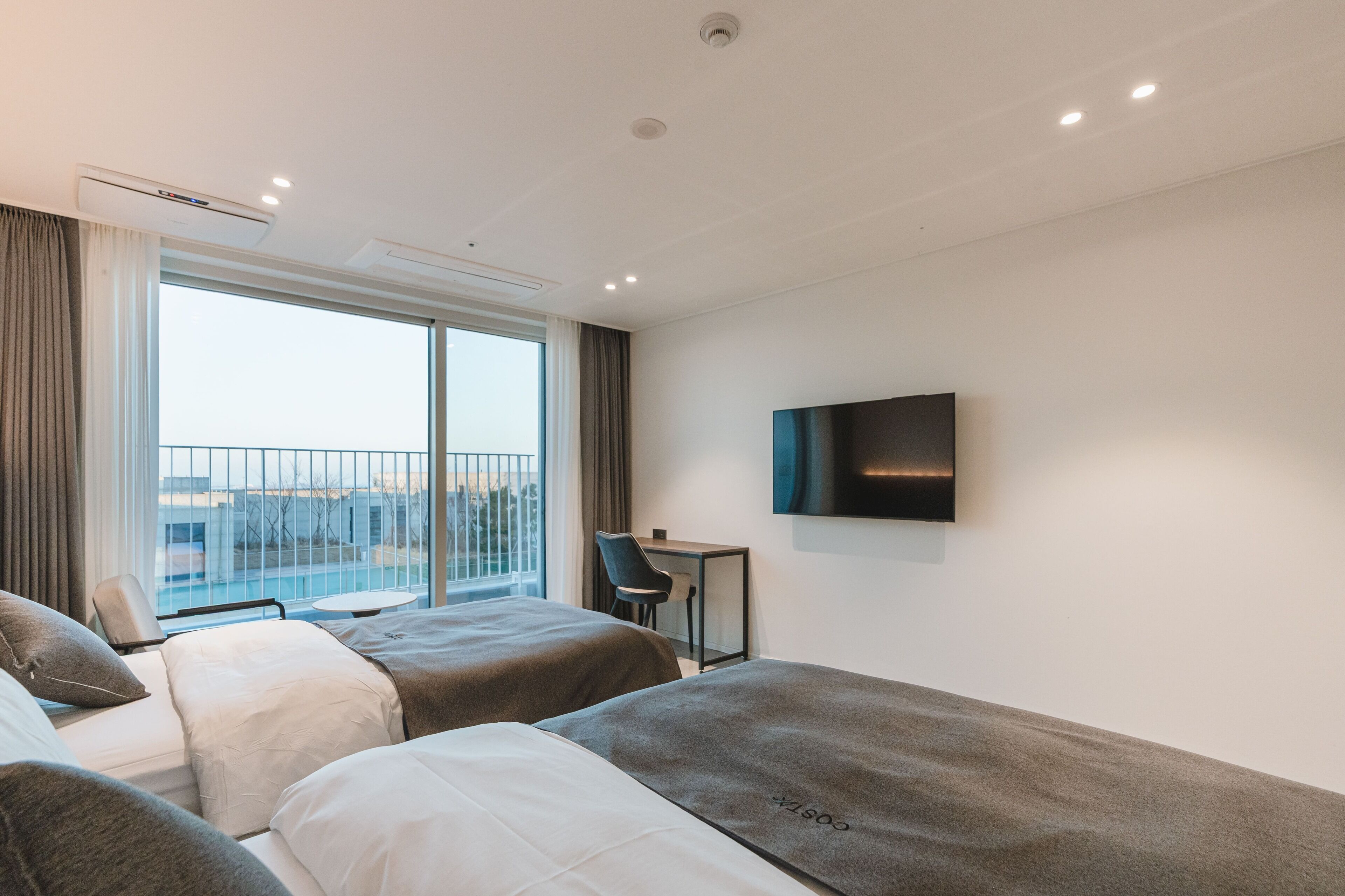 Ein Hotelzimmer mit zwei Betten, einem Schreibtisch, einem Stuhl, einem Fernseher und einem Balkon mit Aussicht.