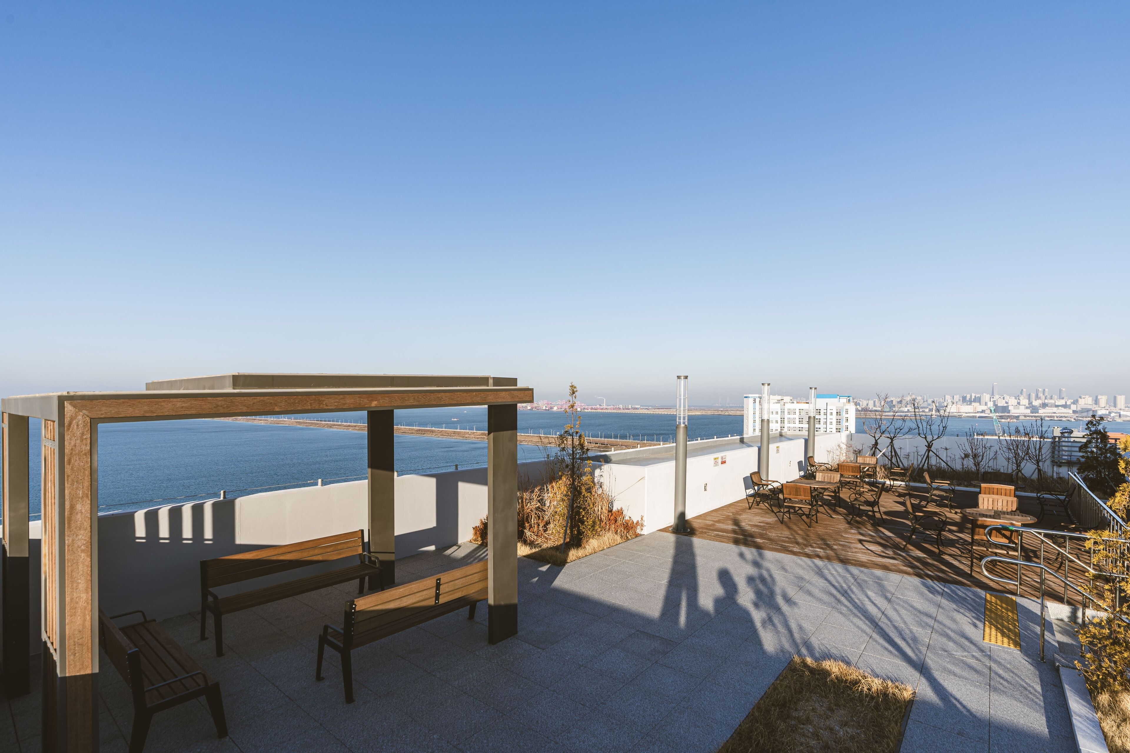 Una terraza al aire libre con vistas al mar y al horizonte de la ciudad, con bancos y mesas de madera.