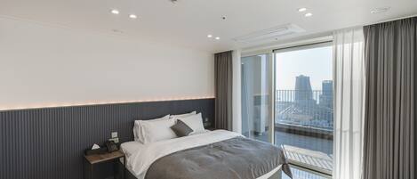 A modern hotel room with a large bed, a bedside table with a phone, and a view of the city through a large window.
