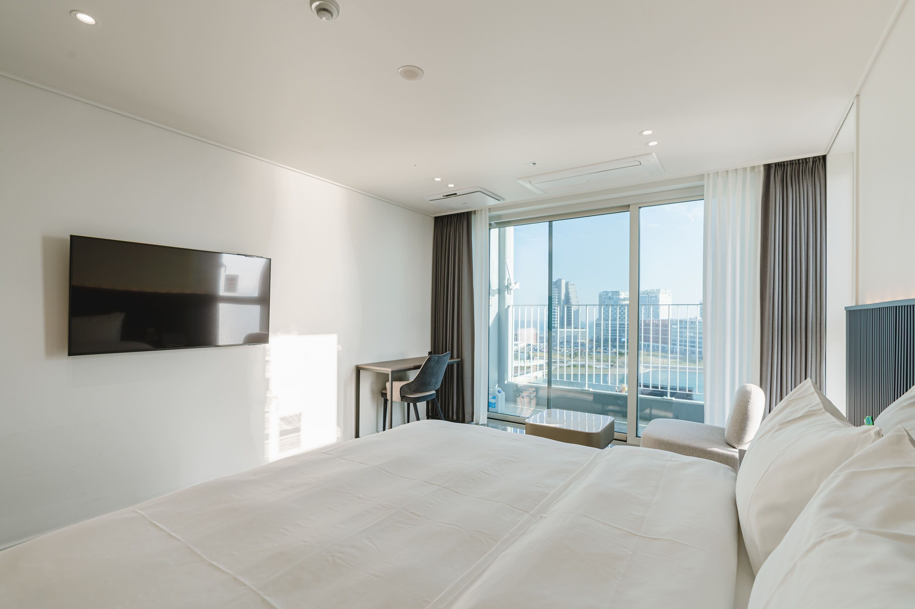 Una habitación de hotel moderna con una cama grande, un escritorio, una silla, un televisor de pantalla plana y vistas a la ciudad a través de amplias ventanas.