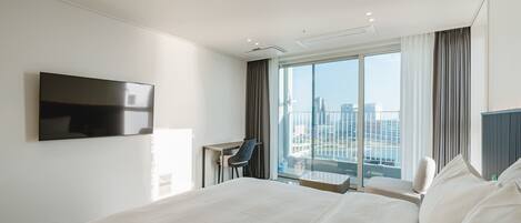 Una habitación de hotel moderna con una cama grande, un escritorio, una silla, un televisor de pantalla plana y vistas a la ciudad a través de amplias ventanas.