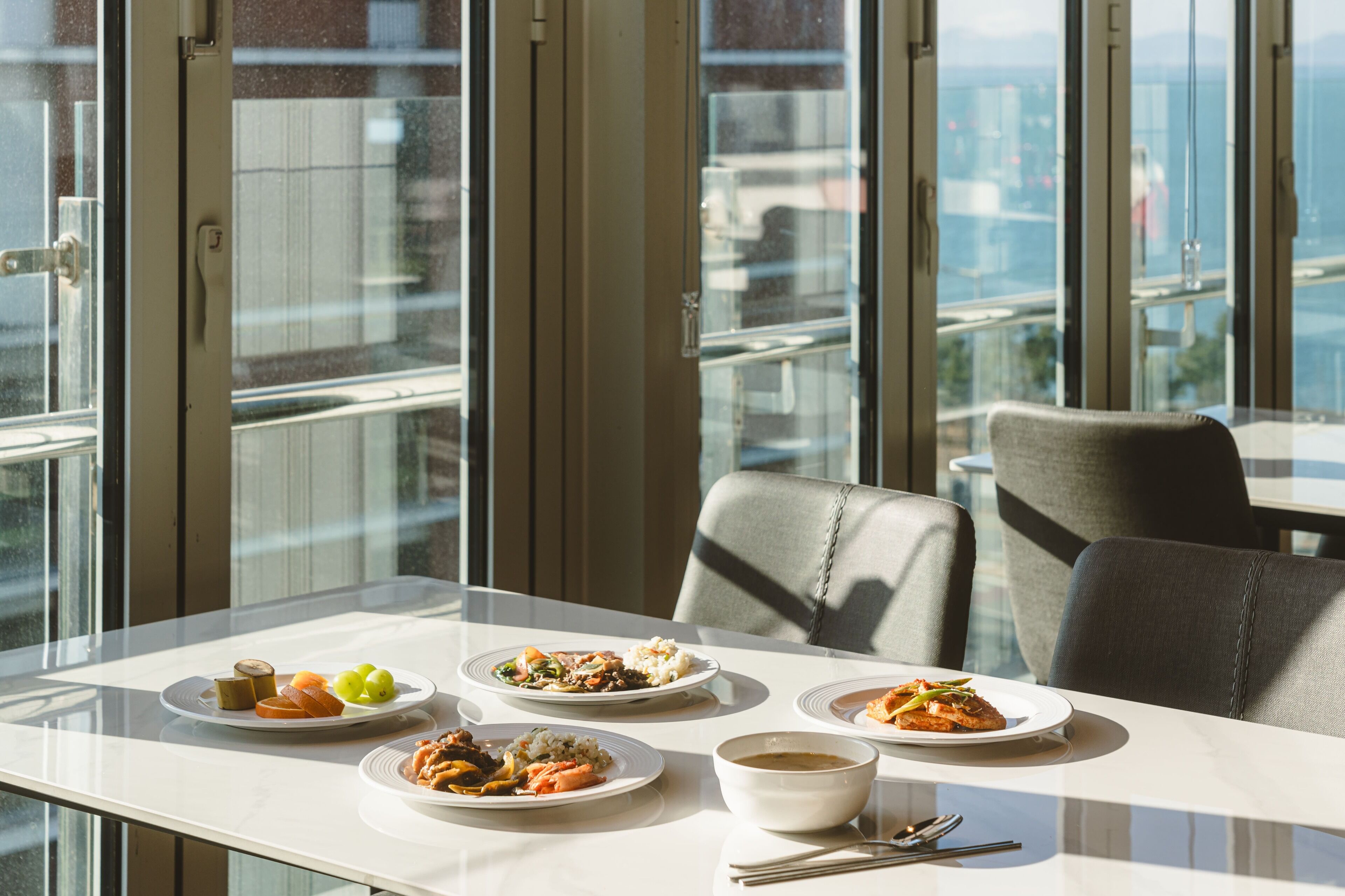 Een eettafel met borden eten en een soepkom in een moderne kamer met grote ramen en uitzicht op de stad.