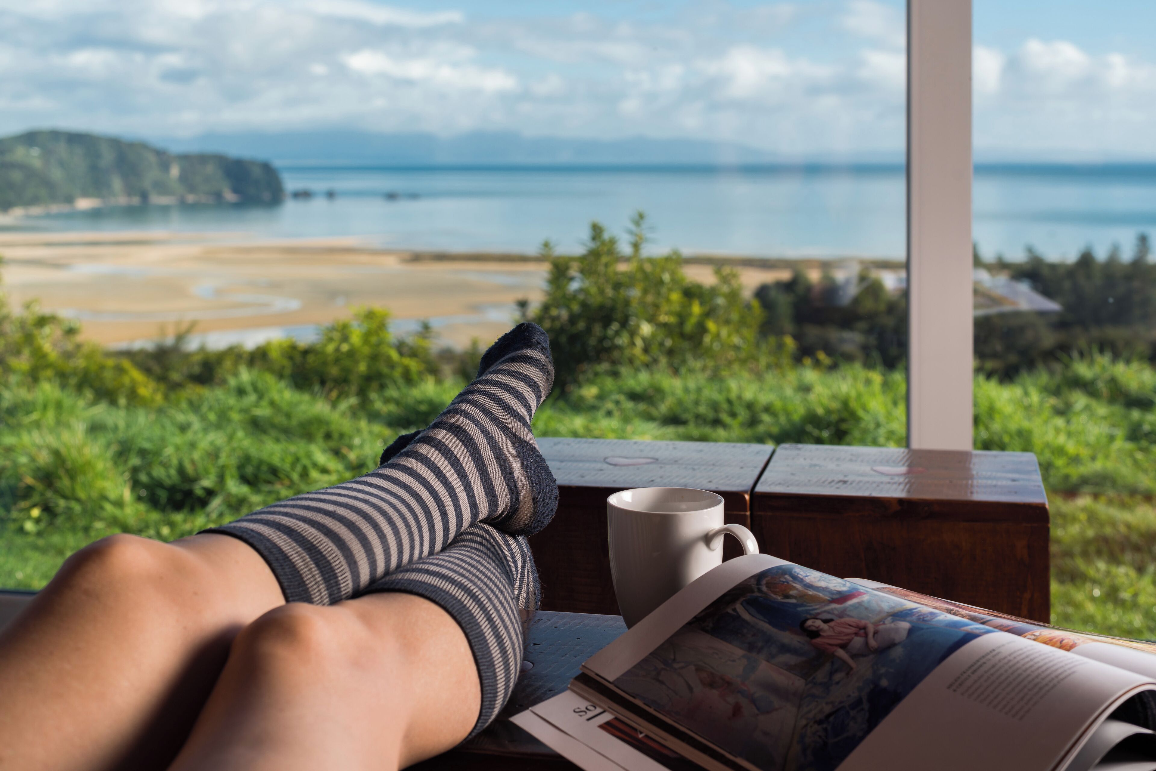 A person relaxing with legs crossed, a book, and a mug on a wooden table, overlooking a beach and ocean view.