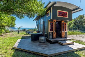 Une petite maison à deux étages, avec une porte et des fenêtres à cadre rouge, située sur une terrasse en bois et offrant une vue sur la mer.
