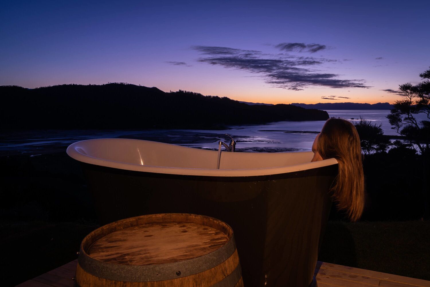 A bathtub with a view of a sunset over water, a wooden barrel, and a person.