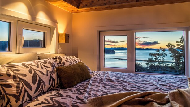 A bedroom with a bed, two windows, and a view of the sea at sunset. - Tiny Greenie - Passive Tiny House, Golden Bay (Takaka)