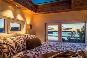 Un dormitorio con una cama, dos ventanas y vista al mar al atardecer.