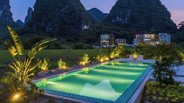A swimming pool with a view of mountains, surrounded by greenery and modern buildings. - Yangshuo 1Q84 Tree Hotel (Guilin)