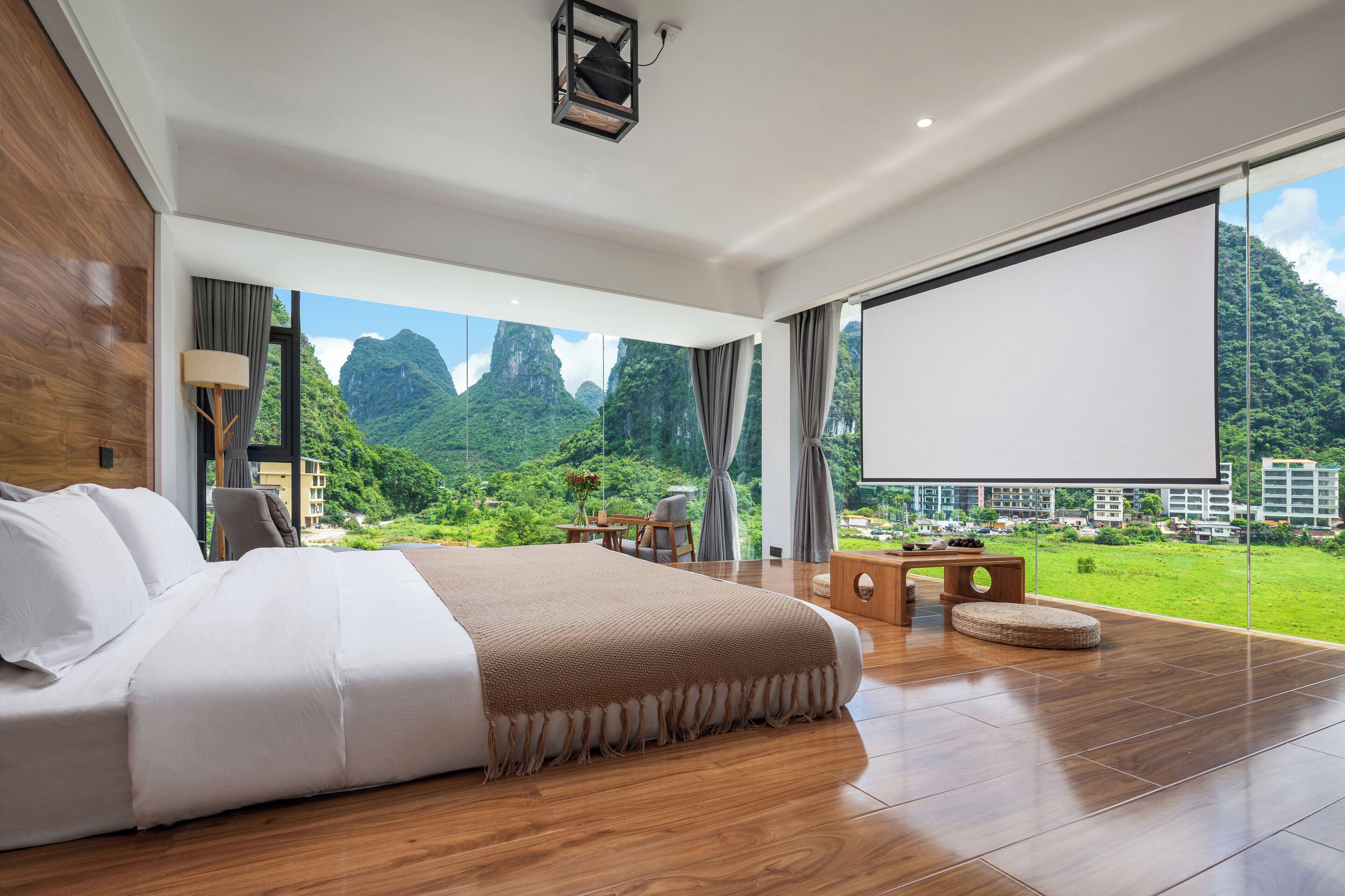 A spacious bedroom with a large bed, a wooden floor, a large window with a view of mountains, and a projector screen.