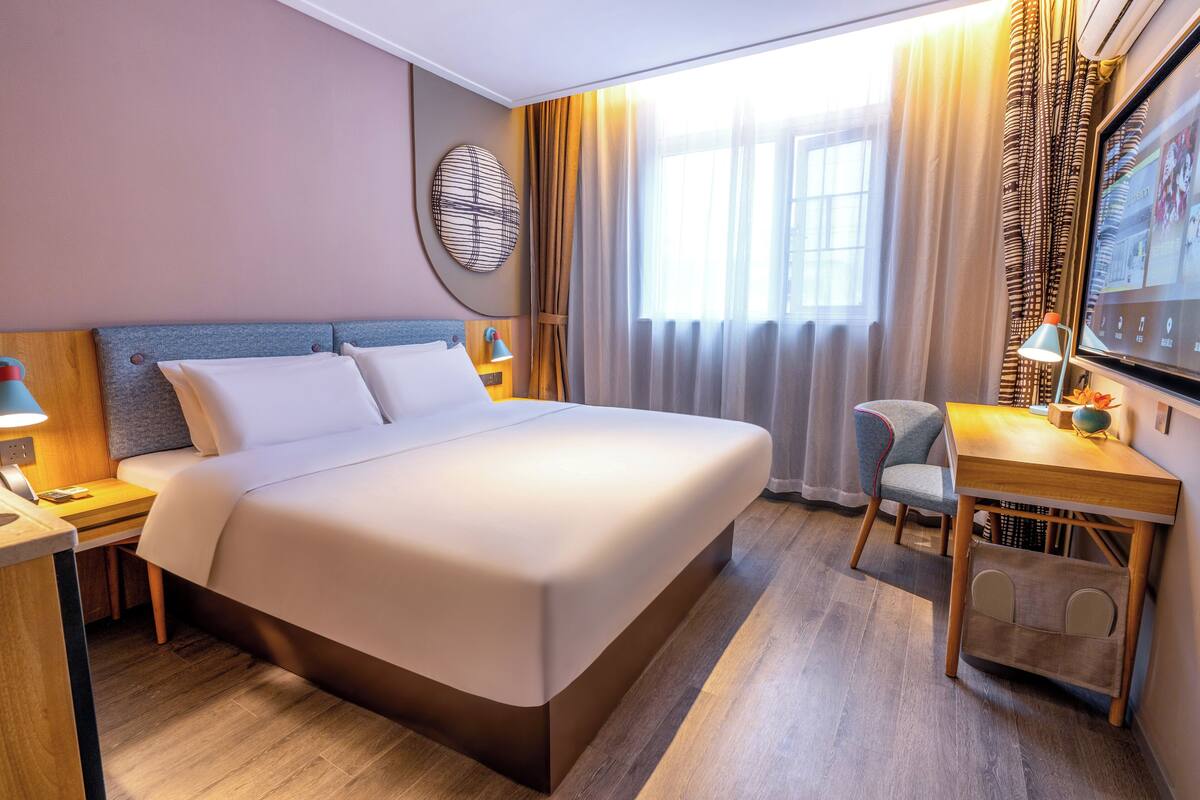 A hotel room with a bed, desk, chair, and a large window with curtains.